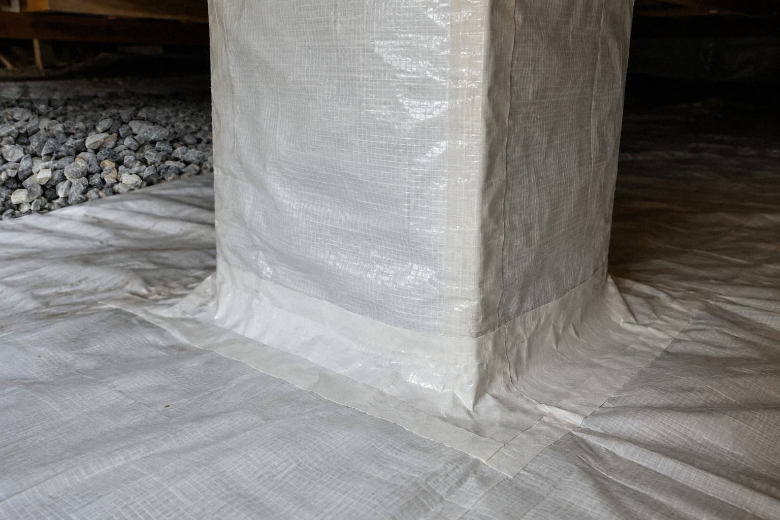 A rendering of the base of a wrapped foundation pier, its vertical seam and the flap where the wrap meets the floor sheet both sealed with tape, gravel visible at the edge of the frame