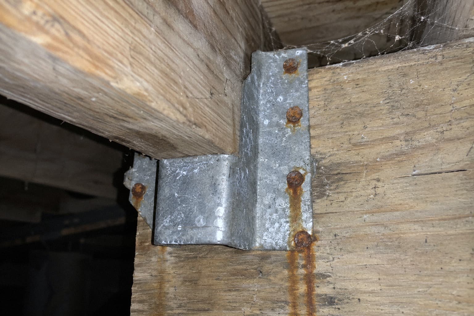 A rendering of a galvanized joist hanger with rust streaking down the wood from every nail head, in a crawlspace with no ground cover