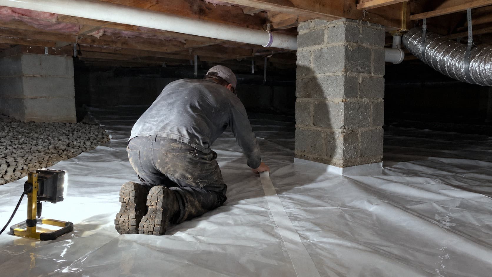 A rendering of a technician seen from behind, kneeling on a new vapor barrier floor and pressing seaming tape down a seam that runs toward a wrapped pier